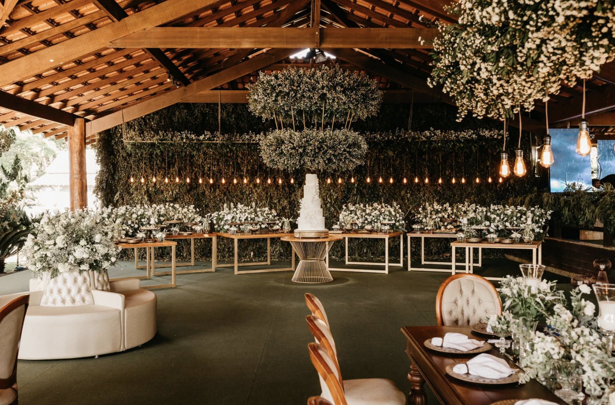 A white tiered wedding cake beneath suspended floral installations, with a living greenery wall, Edison bulbs, and a tufted lounge under a timber pavilion